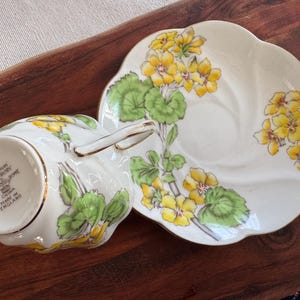 May include: A white teacup and saucer set with a yellow and green floral pattern. The saucer has a scalloped edge. The teacup has a gold rim and the text "GERANIUM SALISBURY FINE CHINA MADE IN ENGLAND" on the bottom.