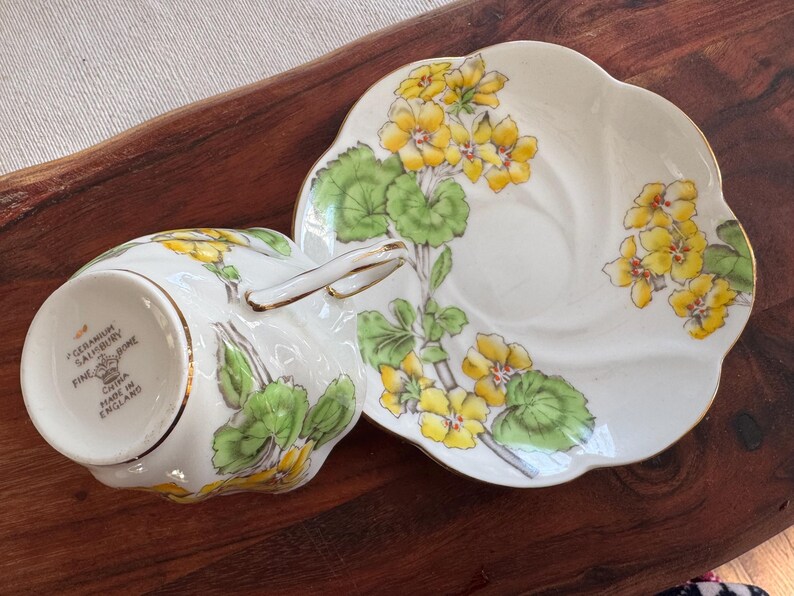 May include: A white teacup and saucer set with a yellow and green floral pattern. The saucer has a scalloped edge. The teacup has a gold rim and the text "GERANIUM SALISBURY FINE CHINA MADE IN ENGLAND" on the bottom.