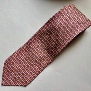 Puede incluir: Una corbata con un patr&oacute;n repetitivo de peque&ntilde;os iconos de anclas de color dorado sobre un fondo morado intenso. La corbata tiene una punta afilada y una textura tejida suave, mostrando una disposici&oacute;n geom&eacute;trica de motivos n&aacute;uticos.