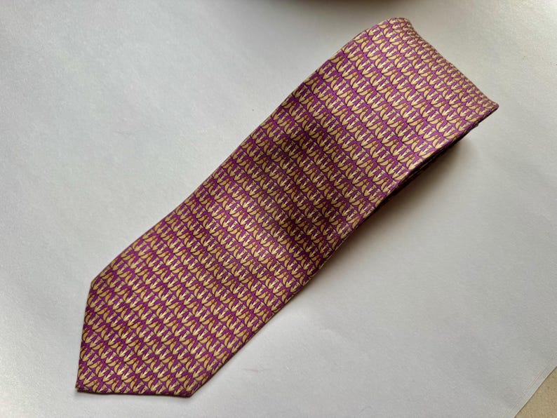 Puede incluir: Una corbata con un patr&oacute;n repetitivo de peque&ntilde;os iconos de anclas de color dorado sobre un fondo morado intenso. La corbata tiene una punta afilada y una textura tejida suave, mostrando una disposici&oacute;n geom&eacute;trica de motivos n&aacute;uticos.