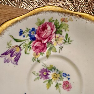 May include: A white porcelain saucer with a gold rim and a floral design. The flowers are pink, purple, yellow, and blue.