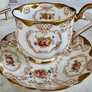 Peut inclure: Un service à thé blanc avec des bordures dorées et des motifs floraux. La tasse et la soucoupe présentent des roses roses et rouges, des accents bleus et des détails dorés ornés. L'ensemble est conçu pour le service du thé.