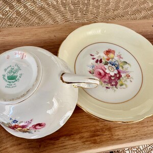 May include: A vintage teacup and saucer set with a floral design. The saucer is a pale yellow color with a gold rim. The teacup has a white background with a floral design in shades of pink, red, and yellow. The bottom of the teacup has a green and white label that reads "Bone China, Handpainted, Made in England".