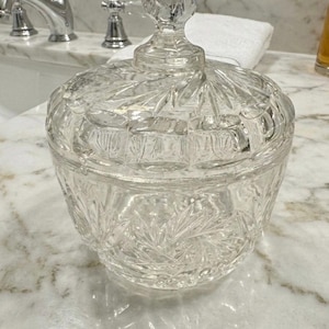 May include: A clear glass candy dish with a matching lid, featuring intricate starburst and geometric cut patterns. The container has a rounded body and a faceted knob handle on top, showcasing a reflective, transparent finish throughout the piece.