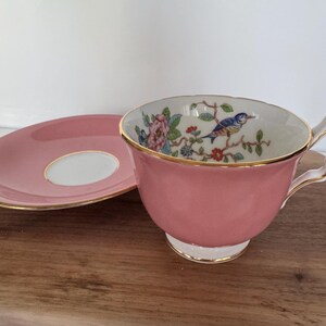 May include: A pink teacup and saucer set with gold trim. The inside of the teacup features a floral and bird design. The saucer is pink with a white center. The set is placed on a wooden surface.