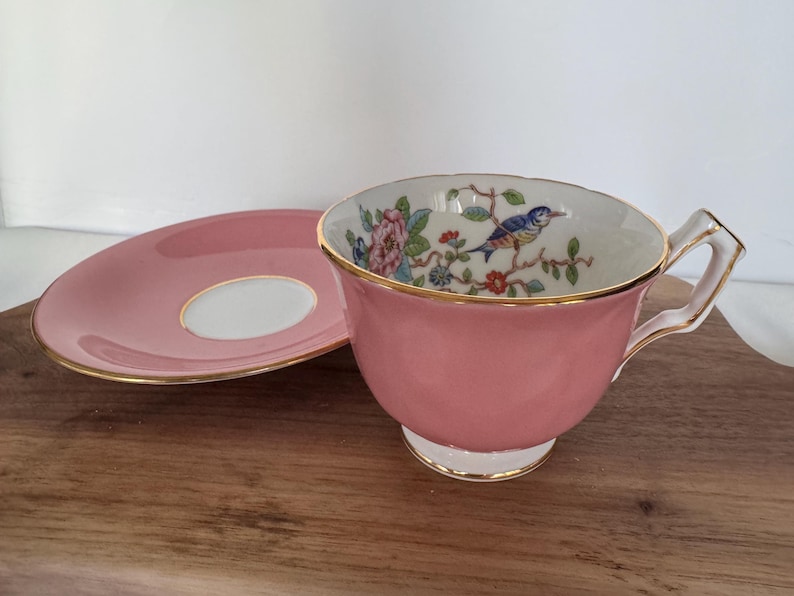 May include: A pink teacup and saucer set with gold trim. The inside of the teacup features a floral and bird design. The saucer is pink with a white center. The set is placed on a wooden surface.
