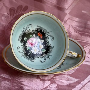 May include: A pale mint green porcelain teacup and saucer set featuring a central floral motif of pink roses and white daisies set against a black scalloped background. The rim of the cup and saucer is trimmed with a thin gold band and scalloped edges.