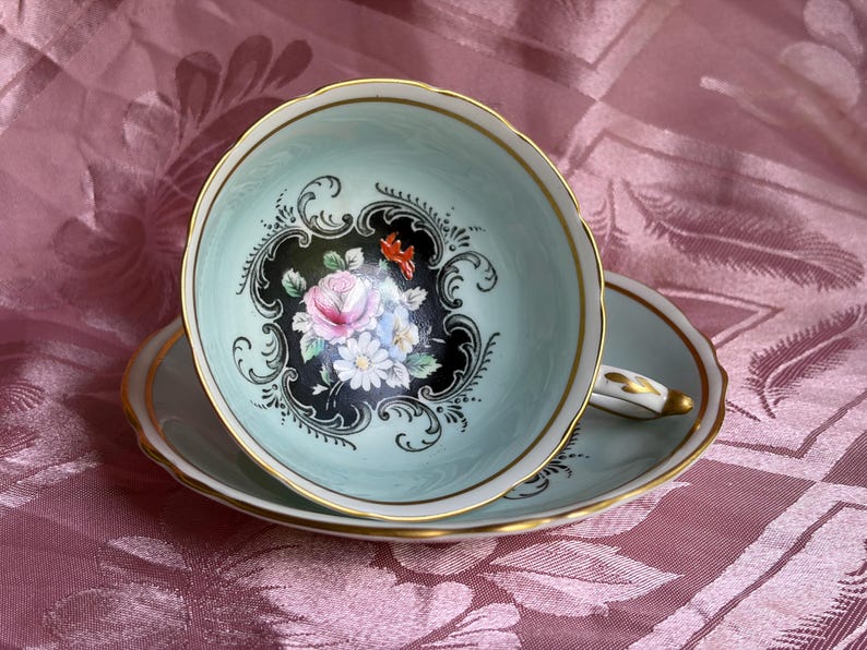 May include: A pale mint green porcelain teacup and saucer set featuring a central floral motif of pink roses and white daisies set against a black scalloped background. The rim of the cup and saucer is trimmed with a thin gold band and scalloped edges.