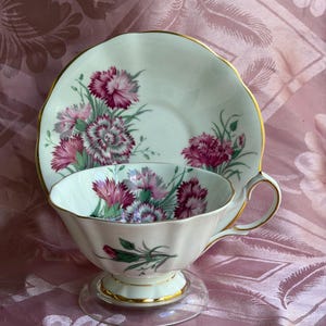 May include: A white porcelain teacup and matching saucer set featuring a scalloped edge with gold trim. The set is decorated with detailed illustrations of pink and magenta carnations with green foliage on both the cup and the saucer.