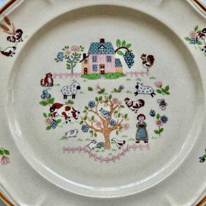 May include: Cream-colored decorative plate with a brown rim and scalloped edges. The center features a pastoral scene with a pink house, farm animals, and a tree. Floral and animal motifs decorate the plate's border.