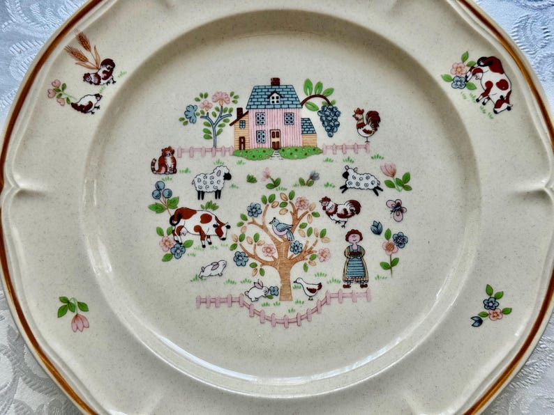 May include: Cream-colored decorative plate with a brown rim and scalloped edges. The center features a pastoral scene with a pink house, farm animals, and a tree. Floral and animal motifs decorate the plate's border.