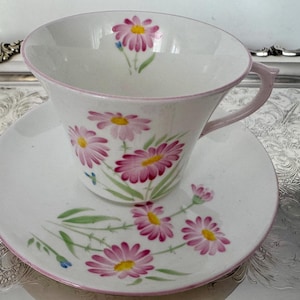 May include: White teacup and saucer set with a pink floral daisy design. The cup and saucer have a pink trim and handle. The daisies have pink petals and yellow centers. The set is on a silver tray.