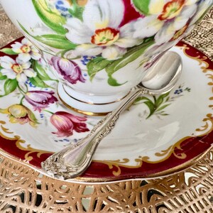 Puede incluir: Juego de taza y platillo de té vintage con diseño floral. La taza y el platillo de porcelana blanca presentan un borde rojo y ribete dorado. Una cuchara de plata descansa sobre el platillo, complementando la estética elegante.