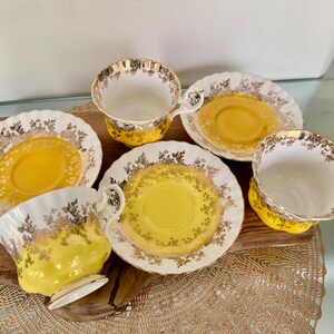 Puede incluir: Un juego de tazas y platillos vintage. Las tazas y los platillos tienen una combinaci&oacute;n de colores blanco y amarillo con adornos dorados. Los platillos tienen un borde festoneado. Las tazas tienen forma redondeada y asa.