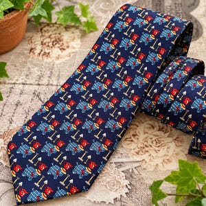 May include: A dark blue necktie featuring a repeating pattern of small red and blue plaid dogs, each accompanied by a miniature garden rake. The tie has a pointed tip and a folded section, showcasing the consistent graphic print across the fabric.