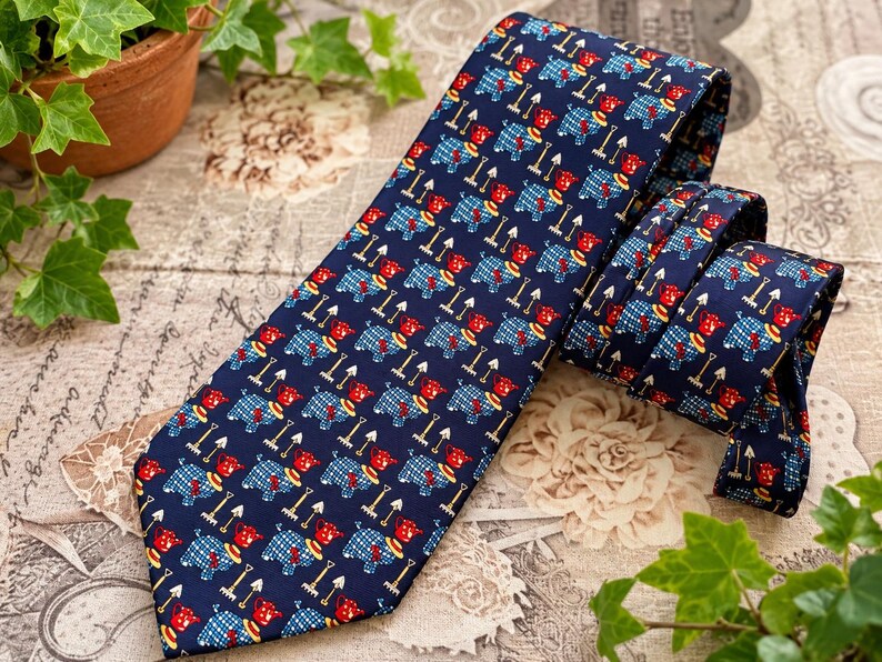 May include: A dark blue necktie featuring a repeating pattern of small red and blue plaid dogs, each accompanied by a miniature garden rake. The tie has a pointed tip and a folded section, showcasing the consistent graphic print across the fabric.