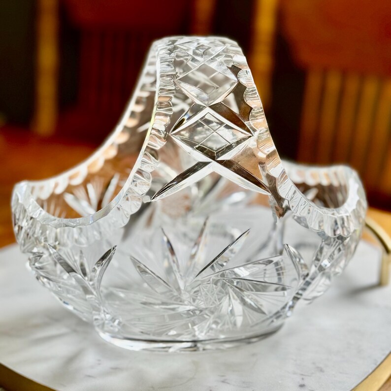 May include: Clear crystal basket with a handle. The basket features intricate starburst and diamond-cut patterns. The handle has a scalloped edge. The basket is designed to hold items and is made of glass.