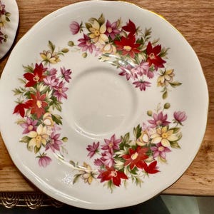 May include: A white porcelain saucer with a gold rim features a floral design with red, pink, and yellow flowers. The saucer has a center well for a teacup.