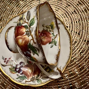 Op de afbeelding: Een wit theekopje en schotel met een gouden rand en een bloemenpatroon met rode, groene en paarse vruchten. Het patroon doet denken aan een vintage ontwerp.