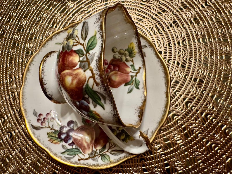 Op de afbeelding: Een wit theekopje en schotel met een gouden rand en een bloemenpatroon met rode, groene en paarse vruchten. Het patroon doet denken aan een vintage ontwerp.