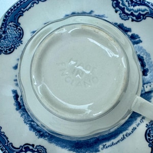 Puede incluir: Una taza de t&eacute; blanca con asa, colocada sobre un platillo a juego. El platillo y la taza presentan un dise&ntilde;o floral azul y blanco con una escena de paisaje. La base de la taza est&aacute; estampada "MADE IN ENGLAND".