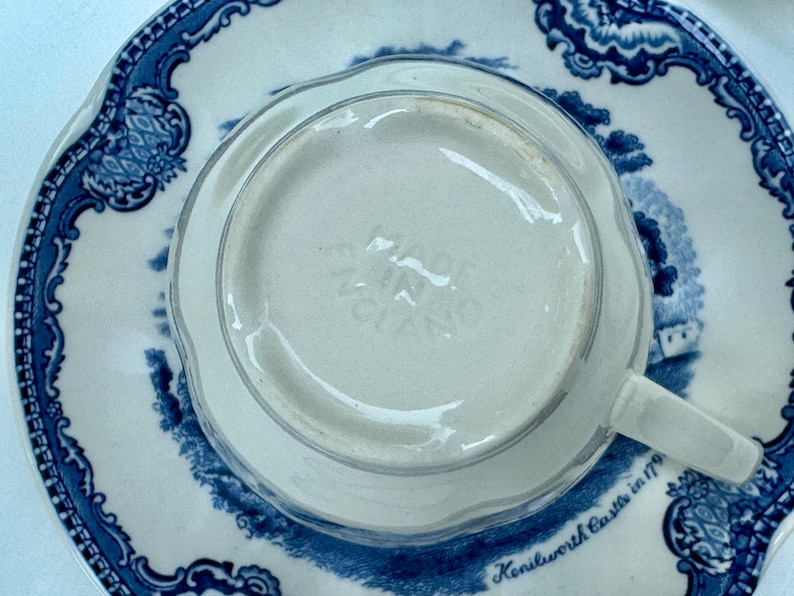 Puede incluir: Una taza de t&eacute; blanca con asa, colocada sobre un platillo a juego. El platillo y la taza presentan un dise&ntilde;o floral azul y blanco con una escena de paisaje. La base de la taza est&aacute; estampada "MADE IN ENGLAND".