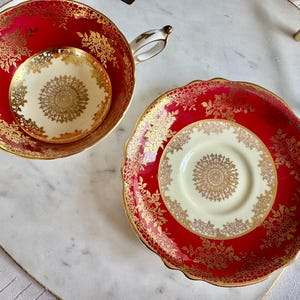 May include: A red teacup and saucer set with gold floral detailing. The teacup and saucer have a cream-colored center with a gold circular design. The set is on a white marble surface.