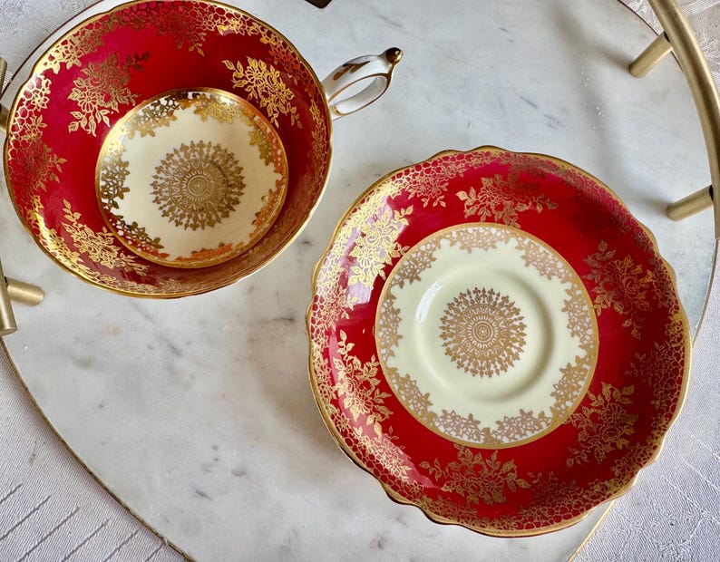 May include: A red teacup and saucer set with gold floral detailing. The teacup and saucer have a cream-colored center with a gold circular design. The set is on a white marble surface.