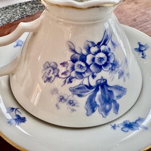 May include: A white porcelain teacup and saucer with a blue floral design. The cup has a delicate handle and a scalloped rim. The saucer has a gold trim.