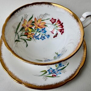 May include: A white teacup and saucer with a gold rim and floral pattern. The cup features a bouquet of yellow, orange, red, and blue flowers. The saucer has a smaller floral design with blue flowers.