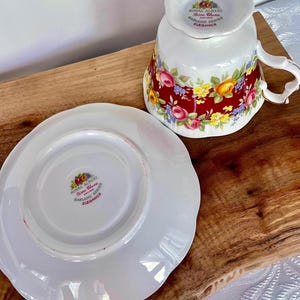 May include: A white teacup and saucer set with a floral design. The teacup is upside down, revealing the Royal Albert Bone China Garland Series Elegance logo. The saucer is white with a scalloped edge. The teacup and saucer are on a wooden surface.