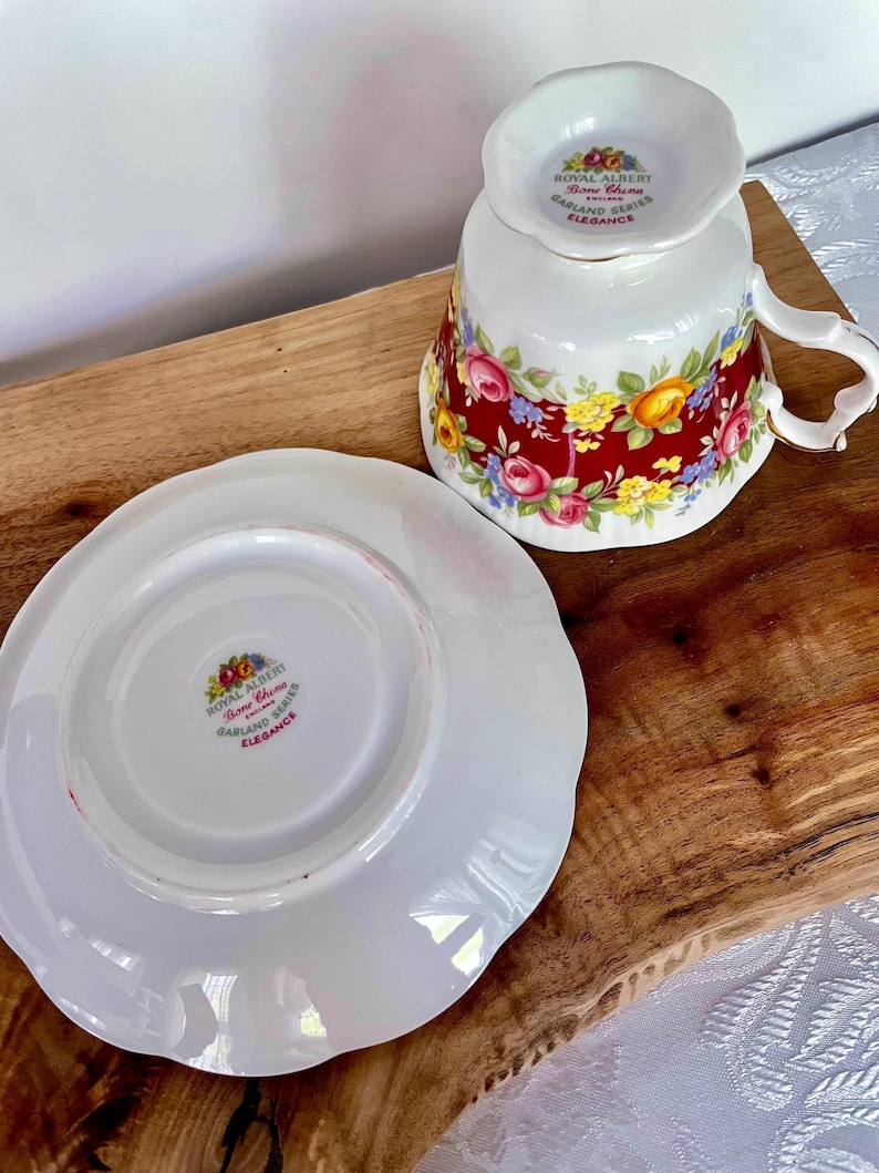May include: A white teacup and saucer set with a floral design. The teacup is upside down, revealing the Royal Albert Bone China Garland Series Elegance logo. The saucer is white with a scalloped edge. The teacup and saucer are on a wooden surface.