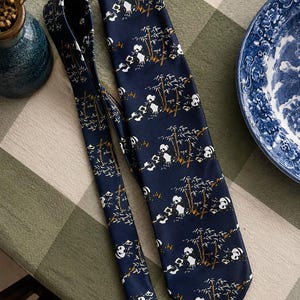 May include: A dark blue necktie featuring a repeating pattern of white and black pandas sitting among brown and white bamboo stalks. The fabric has a smooth, satin-like finish, and the tie is displayed in a folded, overlapping arrangement.