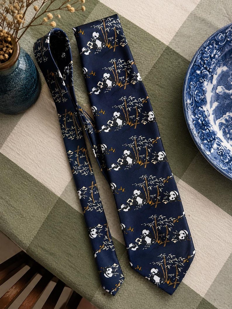 May include: A dark blue necktie featuring a repeating pattern of white and black pandas sitting among brown and white bamboo stalks. The fabric has a smooth, satin-like finish, and the tie is displayed in a folded, overlapping arrangement.
