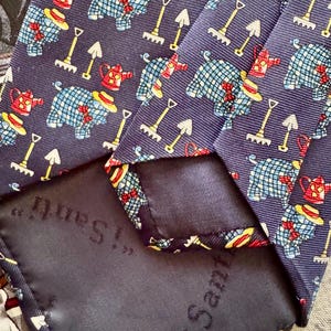 May include: A navy blue necktie featuring a repeating pattern of blue checkered pigs wearing yellow hats and red bow ties, interspersed with red watering cans and yellow garden rakes. The back of the tie features a dark fabric label with the text "i Santi".
