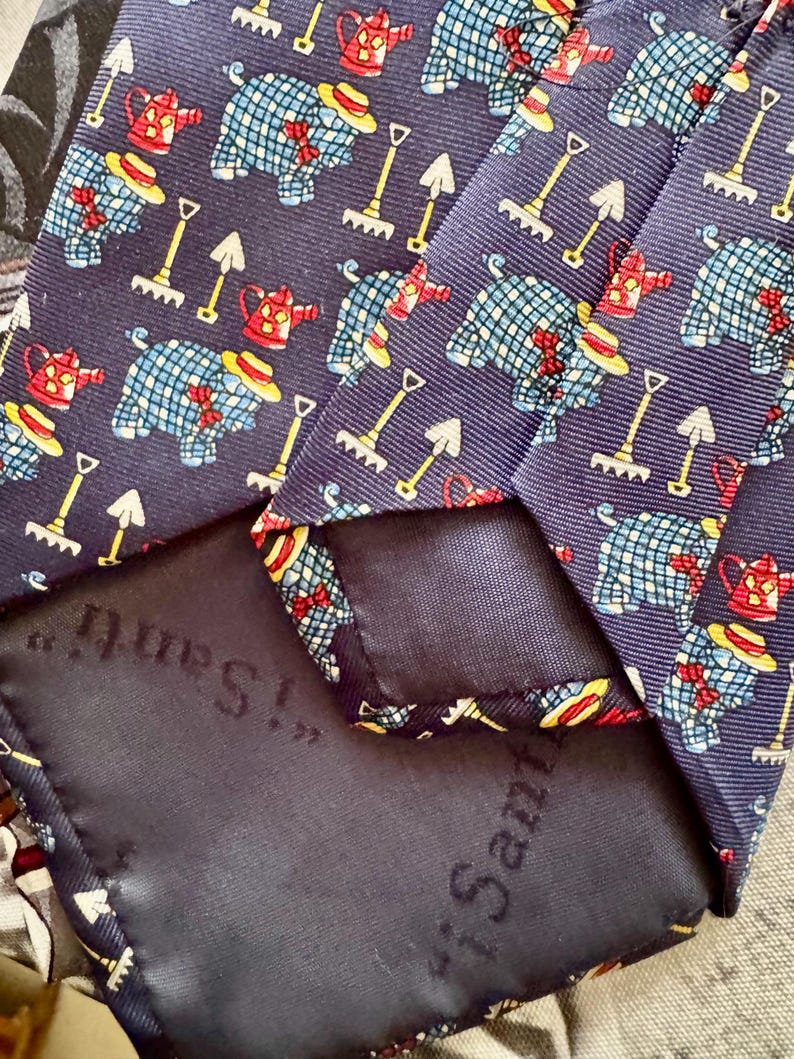 May include: A navy blue necktie featuring a repeating pattern of blue checkered pigs wearing yellow hats and red bow ties, interspersed with red watering cans and yellow garden rakes. The back of the tie features a dark fabric label with the text "i Santi".