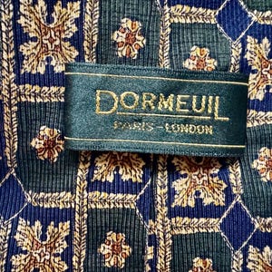 May include: A necktie featuring a repeating geometric pattern of dark green and navy blue squares with gold floral motifs. A dark green fabric label is stitched onto the center, displaying the gold text DORMEUIL PARIS-LONDON.