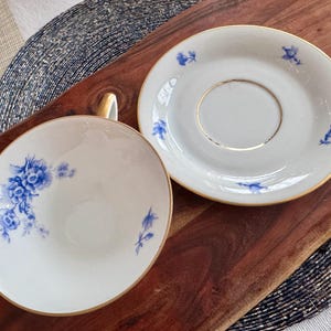 May include: A white teacup and saucer with a blue floral pattern. The saucer has a gold rim.