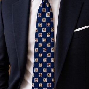 May include: A navy blue necktie featuring a repeating pattern of small, square motifs with red, gold, and white details. The tie is worn with a crisp white dress shirt, a dark navy blue blazer, and a white pocket square in the breast pocket.