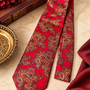 May include: A red necktie featuring a repeating paisley pattern in shades of gold, dark blue, and brown. The fabric has a smooth, satin-like finish, and the tie is displayed in a flat, elongated shape showing the pointed end and the folded tail.