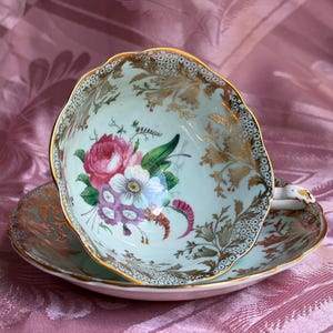 May include: A light mint green porcelain teacup and saucer set featuring a scalloped edge with gold trim. The interior of the cup displays a floral bouquet of pink, white, and purple flowers, accented by intricate gold leaf patterns and stippling.