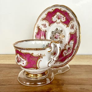 May include: A vintage teacup and saucer set. The white porcelain cup and saucer feature a vibrant pink and gold floral design. The cup has a gold handle and sits on a clear stand. The saucer is placed behind the cup.