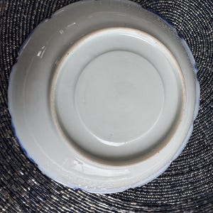 以下が含まれることがあります： 青い縁取りのある白い陶器の皿の裏側。皿は波打った縁取りがあり、花柄で飾られています。