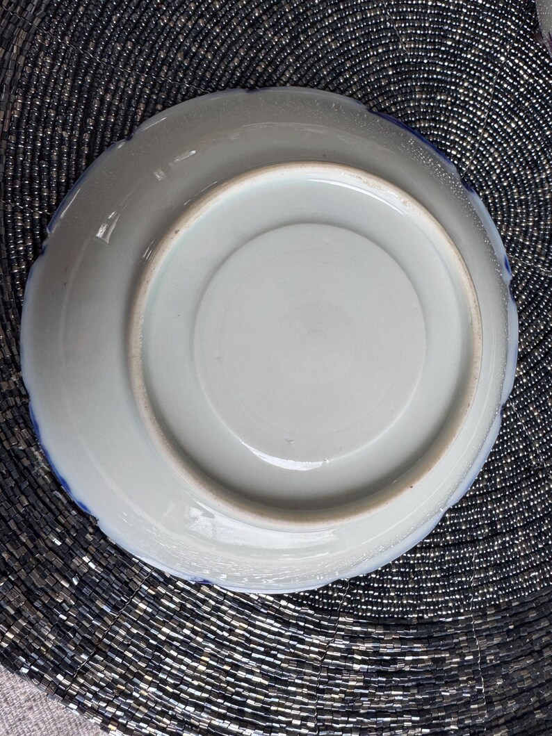 以下が含まれることがあります： 青い縁取りのある白い陶器の皿の裏側。皿は波打った縁取りがあり、花柄で飾られています。