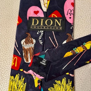 Pu&ograve; includere: Una cravatta blu scuro con un motivo ripetuto di fast food, tra cui patatine fritte, frapp&eacute; e palloncini rosa con cuori. Un'etichetta nera riporta la scritta DION COLLECTION e una piccola etichetta indica il materiale in seta.