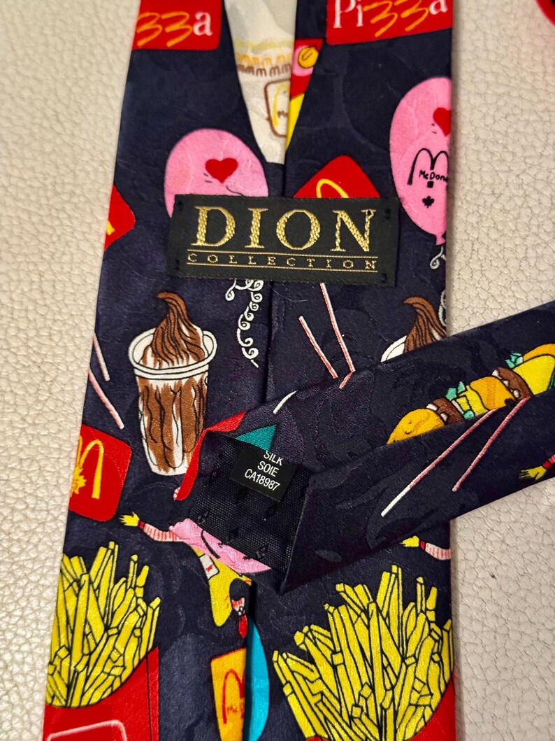 Pu&ograve; includere: Una cravatta blu scuro con un motivo ripetuto di fast food, tra cui patatine fritte, frapp&eacute; e palloncini rosa con cuori. Un'etichetta nera riporta la scritta DION COLLECTION e una piccola etichetta indica il materiale in seta.