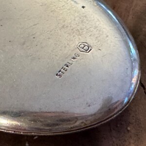 Puede incluir: Primer plano de un objeto plateado de forma ovalada. La palabra "STERLING" est&aacute; estampada en la superficie, junto con un peque&ntilde;o emblema. El objeto tiene una superficie pulida y reflectante, y parece estar hecho de metal. Probablemente una joya o un objeto decorativo.