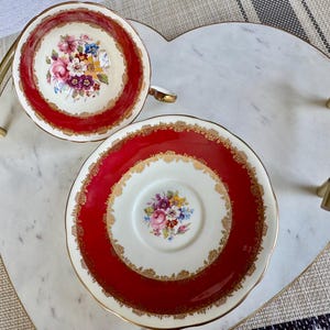 May include: A red and gold teacup and saucer set with a floral design. The saucer has a wide red band with gold trim and a white center with a smaller floral design.