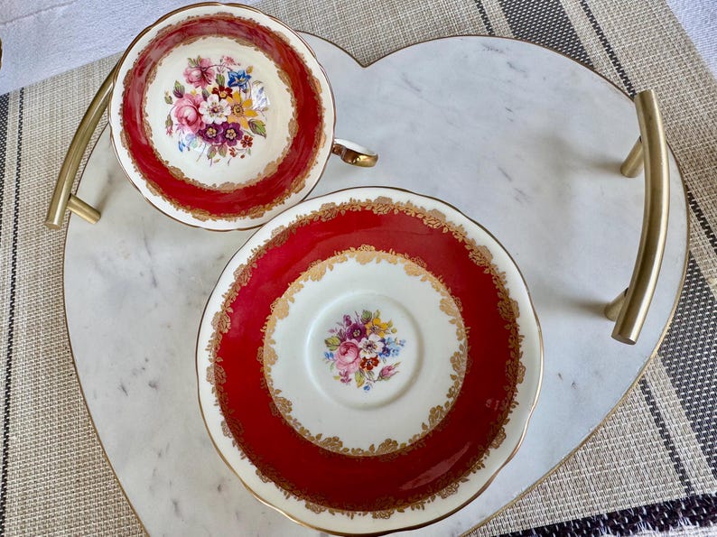 May include: A red and gold teacup and saucer set with a floral design. The saucer has a wide red band with gold trim and a white center with a smaller floral design.