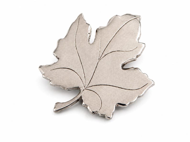 K&ouml;nnte beinhalten: Eine silberfarbene Brosche in Blattform. Die Brosche hat detaillierte Adern und eine strukturierte Oberfl&auml;che. Das Blattdesign ist ein klassisches, von der Natur inspiriertes Accessoire, das sich eignet, jedem Outfit einen Hauch von Eleganz zu verleihen.